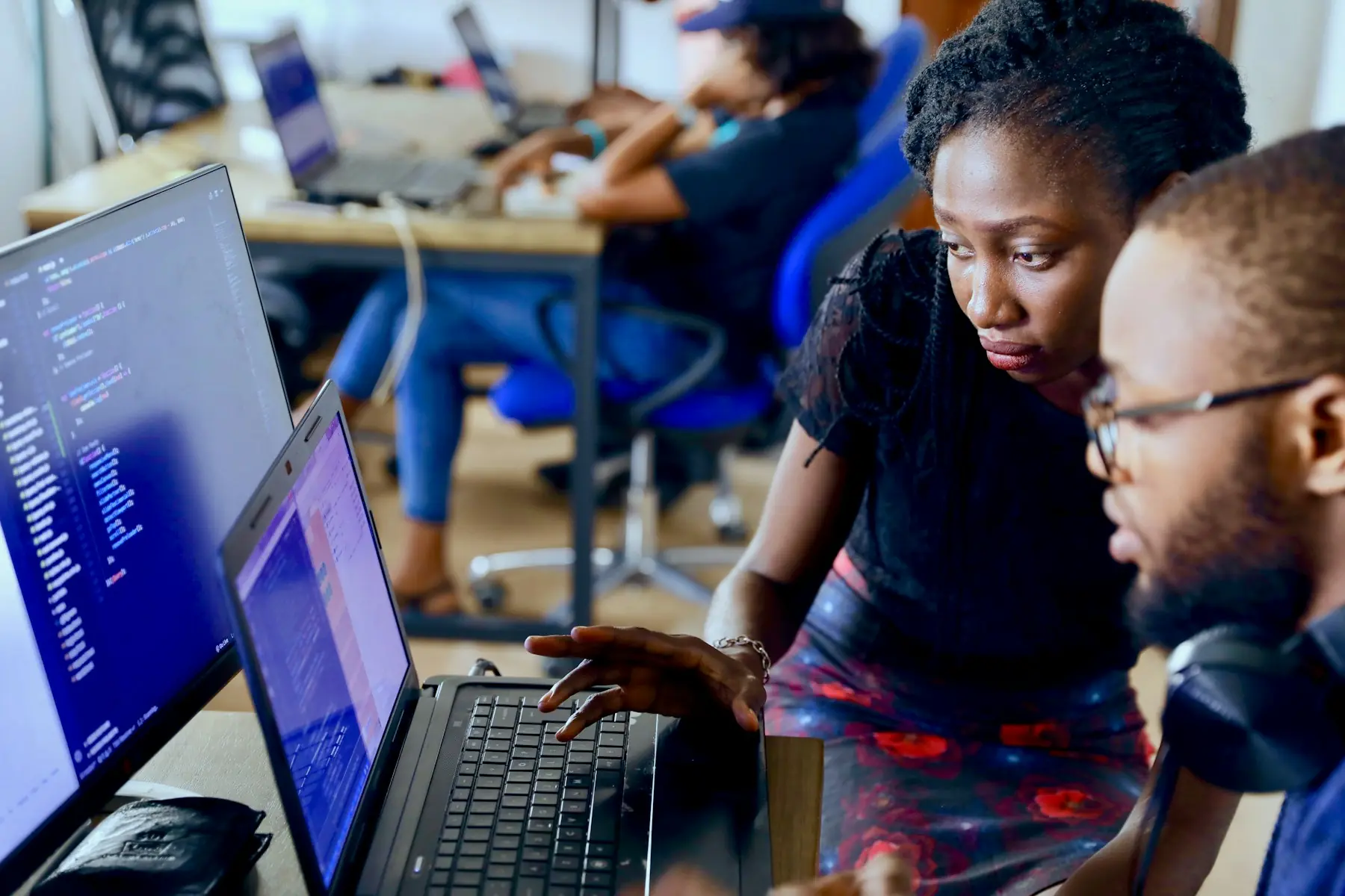 Two young professionals working together at laptops in a busy office